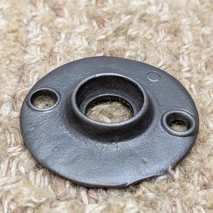 May include: A black metal round plate with a hole in the centre and two smaller holes on the sides. The plate has a smooth, slightly textured surface.