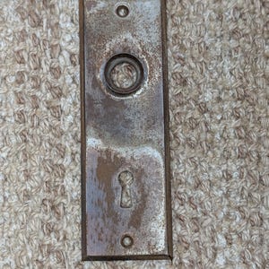 May include: A rectangular metal door plate featuring a weathered, tarnished finish with visible brown rust patches. The plate includes a circular opening at the top for a knob spindle and a lower keyhole opening, with two screw holes at the ends.