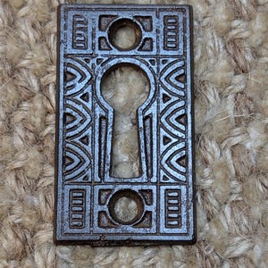 Op de afbeelding: Een sleutelgatbedekking van gietijzer met een ingewikkeld geometrisch ontwerp. De afdekking is donkerbruin en heeft een sleutelgat in het midden.