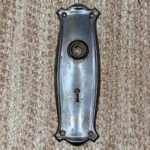 May include: A rectangular metal door plate with a weathered silver finish, featuring a central circular opening for a knob and a lower keyhole. The plate has decorative scalloped edges at the top and bottom, with two small screw holes at each end.