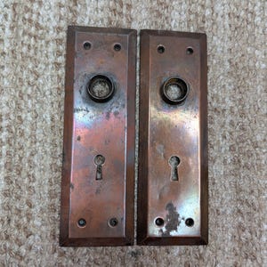 Pode incluir: Duas placas de porta retangulares feitas de cobre envelhecido com uma pátina desgastada. Cada placa apresenta uma abertura circular central para um puxador, uma fechadura abaixo e quatro pequenos orifícios para parafusos nos cantos.