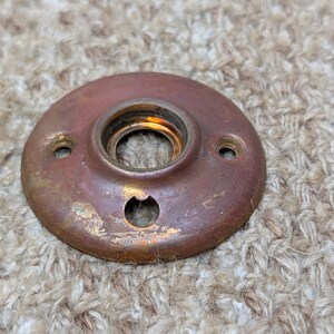 Op de afbeelding: Een ronde deurrozet van koperkleurig metaal met een centrale opening, een metalen binnenrand en drie kleine, gelijkmatig verdeelde schroefgaten langs de rand, die tekenen van oxidatie en oppervlakteslijtage vertoont.