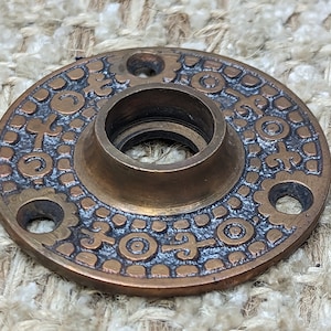 Könnte beinhalten: Eine runde, messingfarbene Türknaufrosette mit einem dekorativen, komplizierten Design. Die Rosette hat ein erhabenes, kreisförmiges Muster mit einem zentralen Loch für den Knauf.