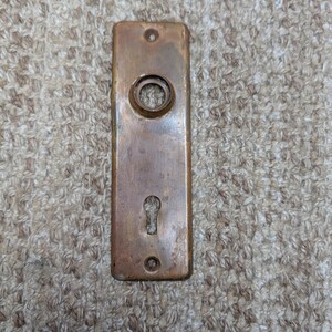 Puede incluir: Una placa de puerta rectangular de latón con una pátina desgastada, que presenta una abertura circular para un pomo de puerta en la parte superior y una abertura para llave en la parte inferior. La placa incluye dos pequeños orificios para tornillos.