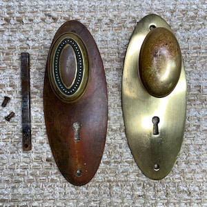 May include: Two antique oval door knobs with keyholes. One knob is brass and the other is brass with a copper patina. Both knobs have a decorative design around the edge.