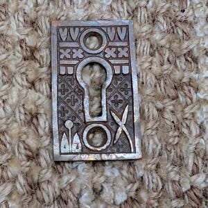 May include: A rectangular, silver-toned keyhole cover with an ornate design. The cover features a keyhole in the center, surrounded by decorative patterns, circles, and cross-like shapes. The metal has a slightly aged appearance.