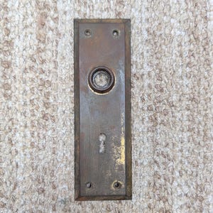 Pode incluir: Uma placa de porta retangular de latão desgastado com uma abertura central circular para maçaneta e uma fechadura inferior. A superfície metálica apresenta sinais de oxidação, pátina e desgaste, com quatro pequenos orifícios para parafusos.