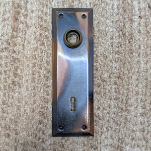 Puede incluir: Una placa de puerta rectangular de metal con acabado en bronce pulido, que presenta una abertura circular para un pomo en la parte superior y una cerradura en la parte inferior. La placa tiene cuatro orificios para tornillos y bordes biselados.