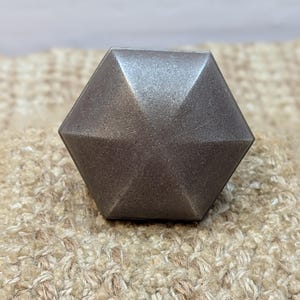 May include: A hexagonal-shaped, dark gray metal knob with a smooth, slightly textured surface.