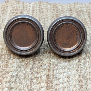 May include: Two round, metal drawer pulls with a copper finish and concentric circles around the edge.