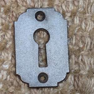 Könnte beinhalten: Ein silberner Schlüsselschlitzabdeckung mit abgerundeter rechteckiger Form und zwei kleinen Löchern auf beiden Seiten des Schlüsselloches.
