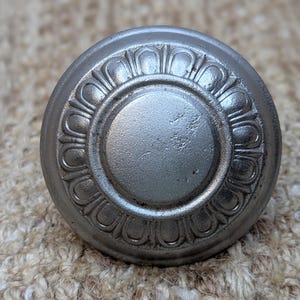 May include: A silver metal knob with a decorative, circular design. The knob has a raised, circular center and a surrounding ring of raised, petal-like shapes.
