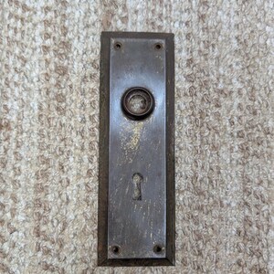 Puede incluir: Una placa de puerta rectangular de metal desgastado con un acabado oscuro y deslustrado con signos visibles de uso. La placa incluye una abertura circular central para un pomo, un orificio para llave en la parte inferior y cuatro agujeros.