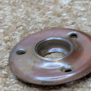 May include: A round, brass-colored metal plate with three holes and a large, circular opening in the center. The plate has a worn, aged appearance.