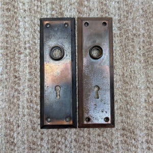 May include: Two rectangular metal door plates with a weathered bronze finish. Each plate features a central circular opening for a knob, a lower keyhole opening, and four small screw holes at the corners. The metal surfaces show signs of oxidation.