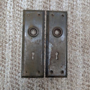 Könnte beinhalten: Zwei rechteckige Türschilder aus Metall mit einer verwitterten, dunkelbronzenen Oberfläche. Jedes Schild verfügt über eine zentrale kreisförmige Öffnung für einen Türknauf, ein Schlüsselloch unten und vier kleine Schraublöcher an den Ecken.