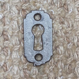 May include: A silver keyhole cover with a decorative design. The cover has two small holes on either side of the keyhole.