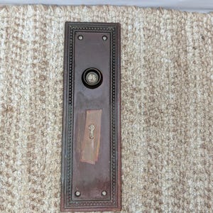 May include: A rectangular, dark brown metal door plate featuring a decorative beaded border, four small screw holes at the corners, a central circular knob mount, and a keyhole opening positioned below the knob mount on the lower half.