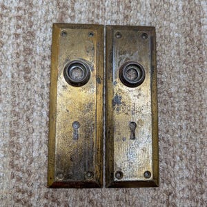 May include: Two rectangular brass door plates with a weathered, tarnished finish. Each plate features a central circular opening for a doorknob, a keyhole below, and four small screw holes at the corners for mounting to a door surface.