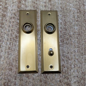 Pode incluir: Duas placas de porta retangulares de latão com acabamento escovado. A placa esquerda apresenta uma abertura circular central. A placa direita inclui uma abertura circular correspondente acima de um pequeno botão de pressão arredondado.