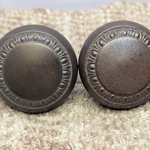May include: Two antique-style door knobs with a dark brown finish. The knobs have a raised, decorative border around the edge.