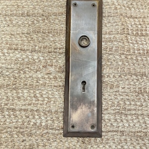 Może przedstawiać: Antyczna, prostokątna, mosiężna tabliczka drzwiowa z dziurką od klucza i okrągłym gałką. Tabliczka ma zużyty, postarzony wygląd i jest lekko matowa.