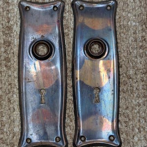 Puede incluir: Dos placas de puerta rectangulares de metal con bordes superior e inferior curvos. Cada placa presenta una abertura circular central para un pomo, una cerradura debajo y cuatro pequeños orificios para tornillos en las esquinas.
