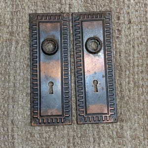 Könnte beinhalten: Zwei antike Türschilder aus Bronze mit Schlüssellöchern und kreisförmigen Grifföffnungen. Die rechteckigen Platten haben einen dekorativen Rand und eine angelaufene Oberfläche. Diese Vintage-Hardware-Teile stammen wahrscheinlich aus einem älteren Haus.