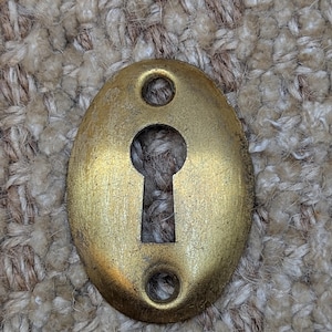 May include: Oval-shaped brass keyhole cover with two small holes on the sides. The keyhole is in the center of the cover.
