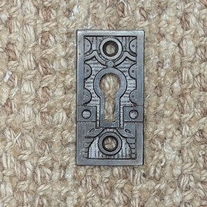 Op de afbeelding: Een zilveren sleutelgatbedekking met een ingewikkeld geometrisch ontwerp. De afdekking is van metaal en heeft een licht verweerde uitstraling.