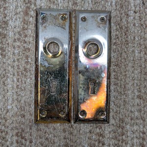 Könnte beinhalten: Zwei rechteckige Türschilder aus poliertem Metall mit silberfarbenem Finish. Jedes Schild verfügt über eine kreisförmige Öffnung für einen Türknauf und eine kleinere Schlüssellochöffnung darunter sowie vier kleine Schraublöcher.