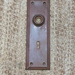 Op de afbeelding: Een rechthoekige, verweerde bruine metalen deurplaat met een centrale ronde knopbehuizing en een sleutelgat eronder. De plaat heeft vier kleine schroefgaten, één in elke hoek, en vertoont over het gehele oppervlak een versleten afwerking.
