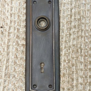 May include: A vintage, dark bronze door plate with a keyhole and a round knob hole. The plate has a decorative border and is slightly curved at the top.