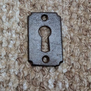 Könnte beinhalten: Rechteckige, antike Metall-Schlüsselplatten mit Schlüsselloch und zwei Schraubenlöchern. Die Platte hat ein dunkles, gealtertes Aussehen, was darauf hindeutet, dass sie aus Eisen oder einem ähnlichen Metall besteht. Das Schlüsselloch ist zentriert.