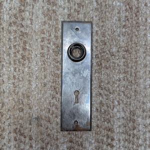 Puede incluir: Una placa de puerta rectangular de metal con un acabado plateado desgastado. La placa presenta una abertura circular para un pomo de puerta en la sección superior y una abertura vertical para llave en la sección inferior, con orificios para tornillos.