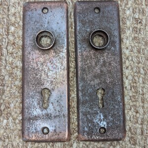 May include: Two antique door plates made of metal with a rusty finish. Each plate has a keyhole and a round hole in the centre.