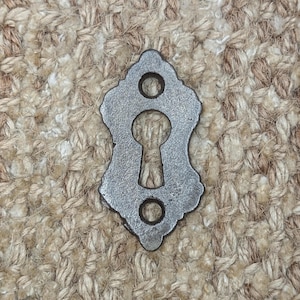 Op de afbeelding: Een zilveren sleutelgatbedekking met een decoratief, sierlijk ontwerp. De afdekking heeft twee kleine gaten aan weerszijden van het sleutelgat.