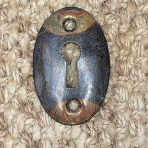 Op de afbeelding: Ovale sleutelgatbedekking van metaal met een versleten, verweerde afwerking. De afdekking is donker zilverkleurig met hints van messing. Het heeft twee kleine gaten aan weerszijden van het sleutelgat.