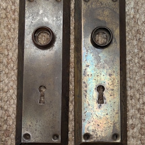 May include: Two antique rectangular door plates with keyholes and circular knob openings. The plates are made of metal with a tarnished, aged appearance. Each plate has four screw holes and a raised border.