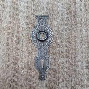 Op de afbeelding: Een verticaal, langwerpig metalen deurschild met een donkergrijze afwerking. Het schild heeft een centrale ronde opening voor een knop en een onderste sleutelgatopening, versierd met symmetrische florale en geometrische reliëfpatronen.