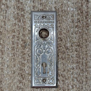 Puede incluir: Una placa de puerta rectangular de metal plateado con un intrincado patrón de volutas en relieve. La placa incluye una abertura circular en la parte superior para el pomo y una cerradura en la parte inferior, con dos pequeños orificios para tornillos.