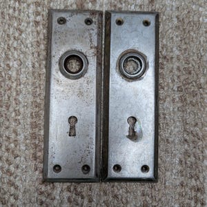 Könnte beinhalten: Zwei rechteckige Türschilder aus Metall mit verwitterter silberner Oberfläche. Jedes Schild verfügt über eine zentrale kreisförmige Öffnung für einen Türknauf, ein Schlüsselloch darunter und vier kleine Schraublöcher an den Ecken.