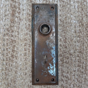 Könnte beinhalten: Eine rechteckige Türschildplatte aus Metall mit einer verwitterten, rostigen Oberfläche und sichtbarer brauner sowie grauer Oxidation. Die Platte verfügt über eine zentrale kreisförmige Öffnung für einen Türknauf, ein Schlüsselloch und vier Schraublöcher.