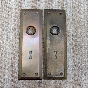 Op de afbeelding: Twee rechthoekige koperen deurplaten met een verweerde patina. Elke plaat heeft een centrale ronde opening voor een deurkrukstift en een onderste sleutelgat. Vier kleine schroefgaten bevinden zich op de hoeken van elke metalen plaat.