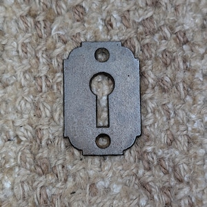 May include: A rectangular metal keyhole escutcheon plate with a dark, weathered finish. The plate features a central keyhole opening shaped like a classic keyhole and two circular screw holes, one positioned above and one below the keyhole.
