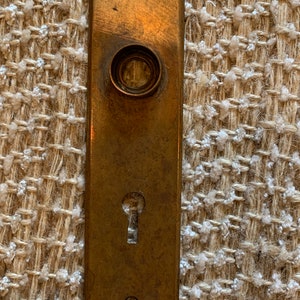 Pode incluir: Uma placa de porta de latão com uma fechadura e um botão redondo. A placa tem um aspecto desgastado e antigo.