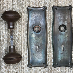 May include: Two antique door plates with a keyhole and a round knob hole, and a matching doorknob with a round top and a metal shaft. The plates and knob are a dark, aged metal with a blue-green patina.