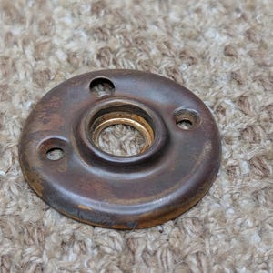 Könnte beinhalten: Eine kreisförmige Türrosette aus dunkelbraunem Metall mit einer verwitterten, gealterten Oberfläche. Das Beschlagteil hat eine zentrale Öffnung mit einem messingfarbenen Innenring und drei kleinen, gleichmäßig verteilten Schraublöchern.