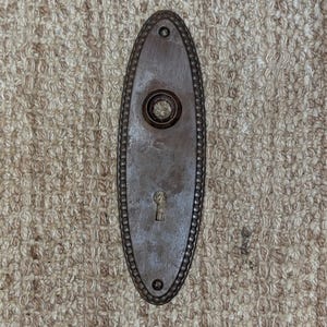 May include: An oval-shaped metal door plate with a dark brown finish. The plate features a beaded border, a central circular opening for a door knob, and a keyhole opening below. There are two small screw holes at the top and bottom ends.
