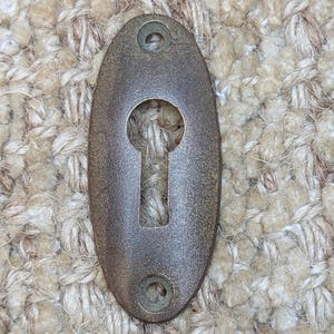 Op de afbeelding: Een vintage ovale sleutelgatbedekking van metaal met een donkerbruine afwerking. De afdekking heeft twee kleine gaten boven en onder voor bevestiging.